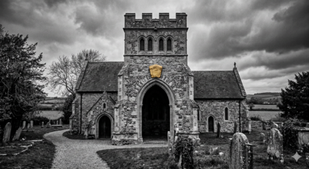 A black and white image of a stone church on a cloudy day. There are trees and grass around the church and a small graveyard in front. The capstone above the arched main entrance is the only thing with color - gold.
